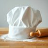 A white chef's hat and rolling pin on a floured counter.
