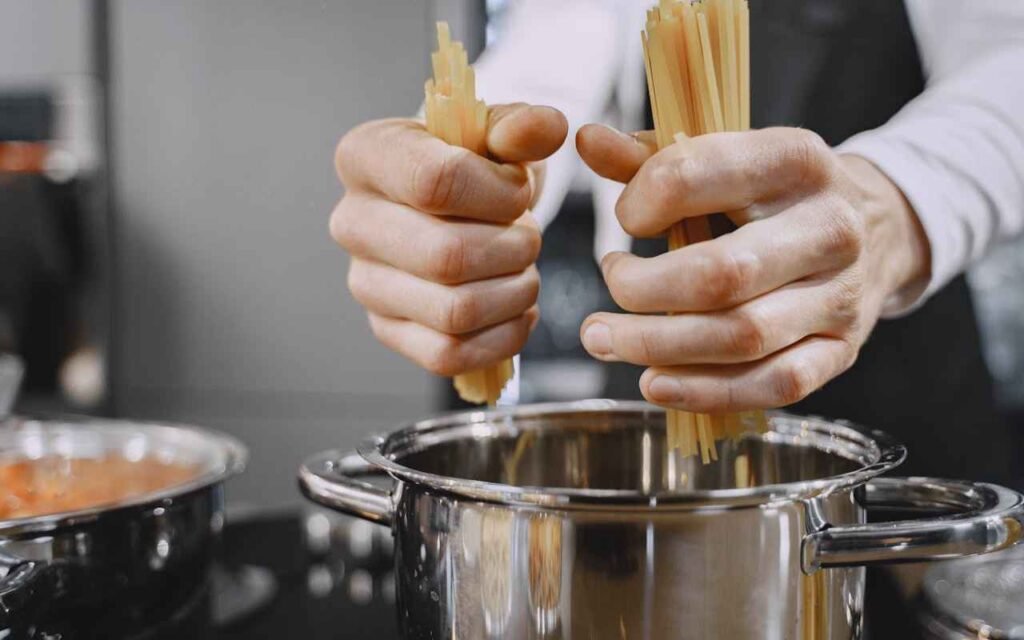 Close-up of hands placing spaghetti pasta into a lightweight stainless steel pot. The shot emphasizes wide, symmetrical loop handles designed to reduce wrist lever-arm torque for home cooks managing hand arthritis.