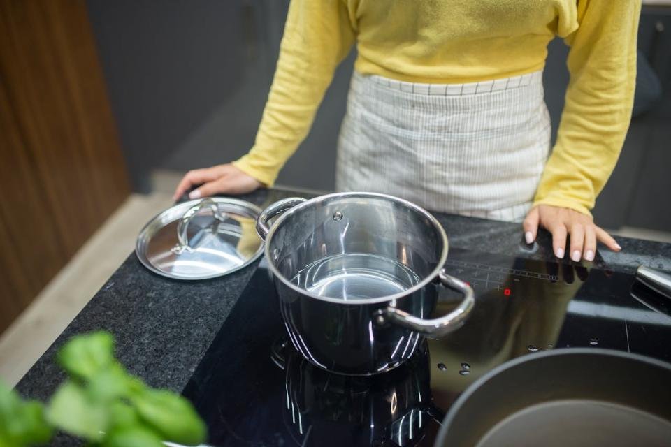 A lightweight thin-gauge stainless steel pot filled with water sitting on a smooth black induction cooktop surface. The shot displays wide dual side-loop handles designed to protect senior wrist joints from excessive lifting strain.