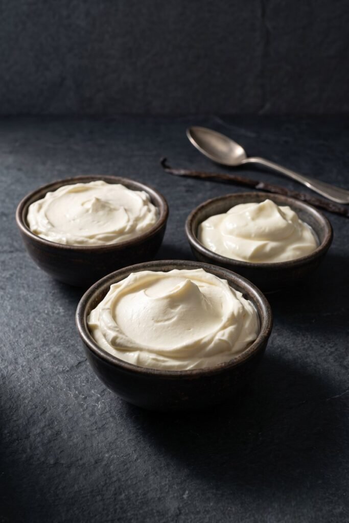 A professional dark food photography shot displaying a bowl of velvety Italian mascarpone cheese next to custom cream cheese and crème fraîche substitutes on a black slate counter.