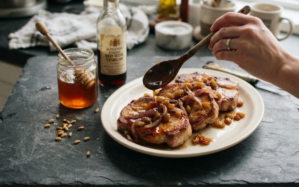 A raw, unedited photo showing a hand using a wooden spoon to drizzle a glossy amber agrodolce sauce with raisins over sliced pork chops on a plate.